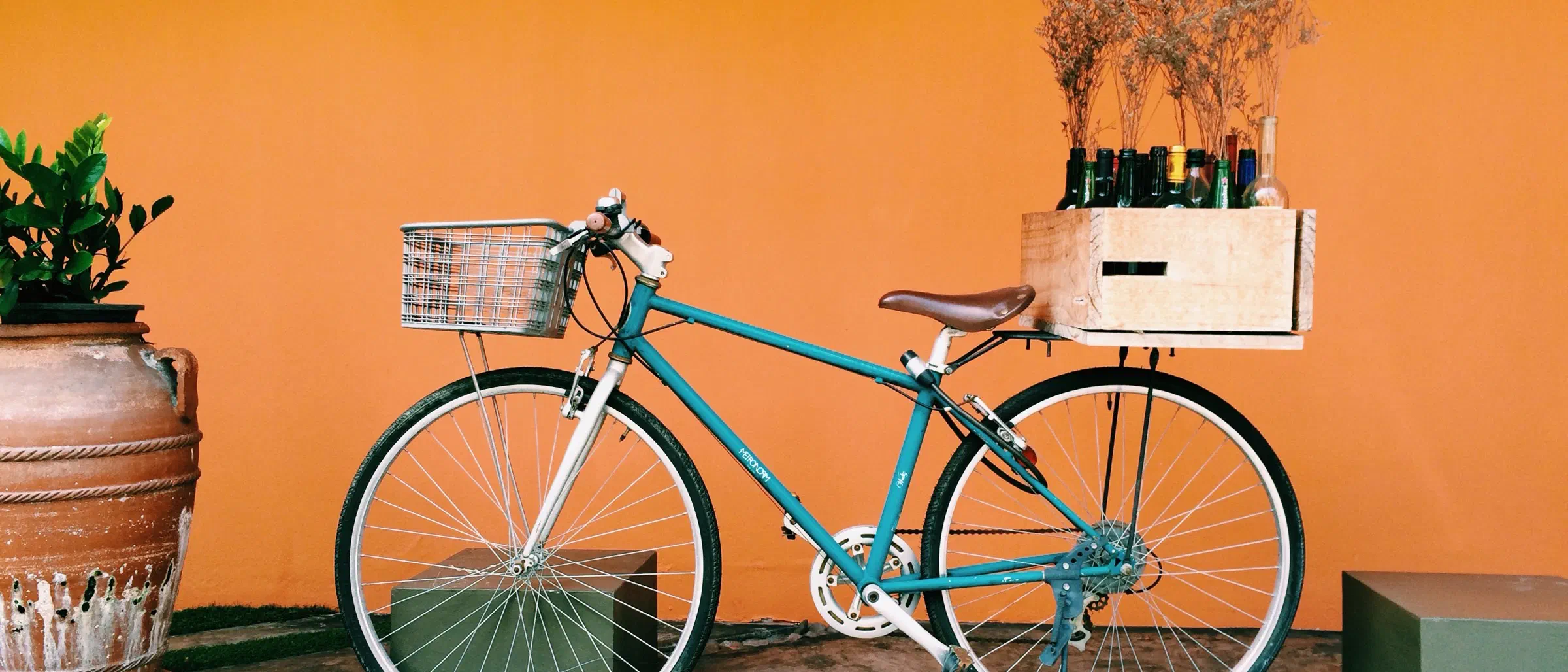 Un vélo de ville bleu canard, avec panier à l'avant et cagette sur le porte-bagages, appuyé contre un mur orange terre cuite.